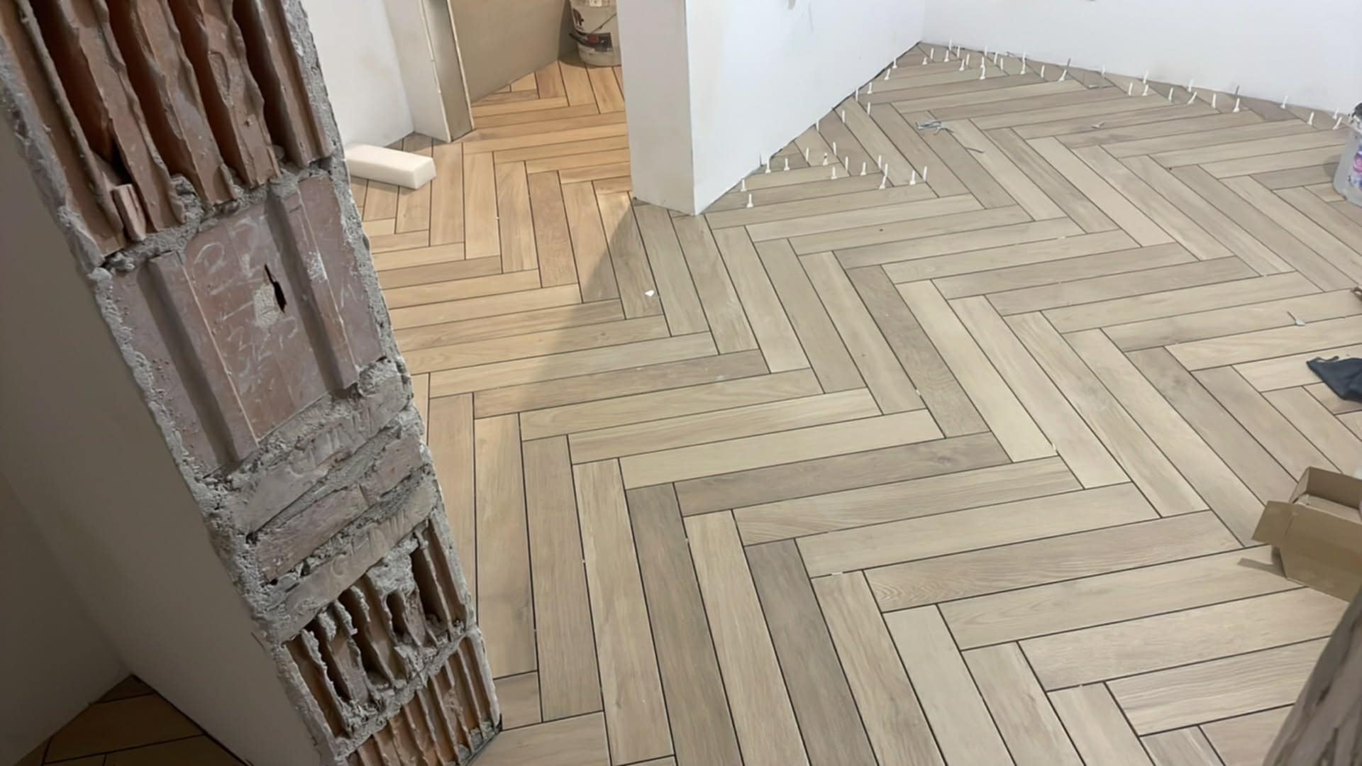 Jasne panele ułożone w jodełkę, widoczne białe krzyżyki dystansowe. Fragment ściany z cegły w surowym stanie. Widok na sąsiednie pomieszczenie.