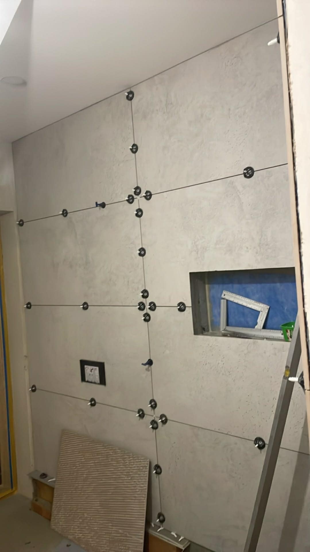 Ściana z szarymi płytkami, system poziomowania płytek z czarnymi klipsami i sznurkami, wnęka z niebieskim tłem, płytka pod ścianą. Widok na etap prac wykończeniowych.