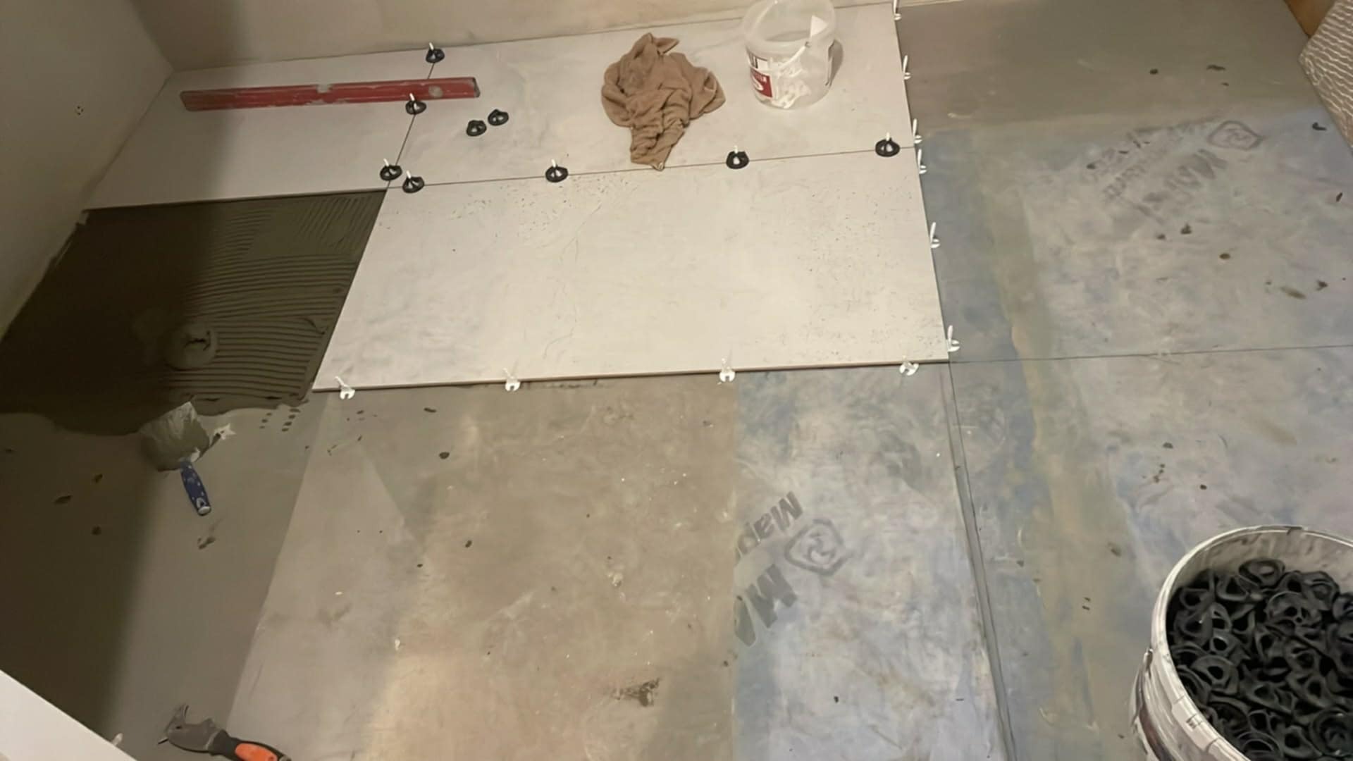 Układanie płytek podłogowych z użyciem poziomicy i systemu poziomowania. Częściowo ułożone płytki z widoczną warstwą kleju. Wiadro z klipsami w rogu.