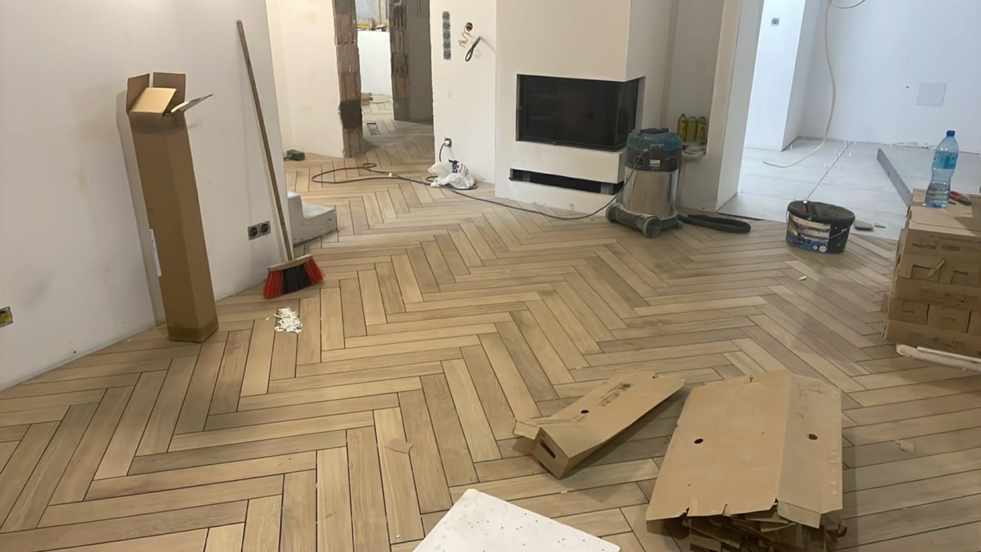 Jasne panele ułożone w jodełkę francuską w przestronnym wnętrzu. Widoczne kartony, odkurzacz i akcesoria malarskie. Częściowo wykończone ściany i kominek.