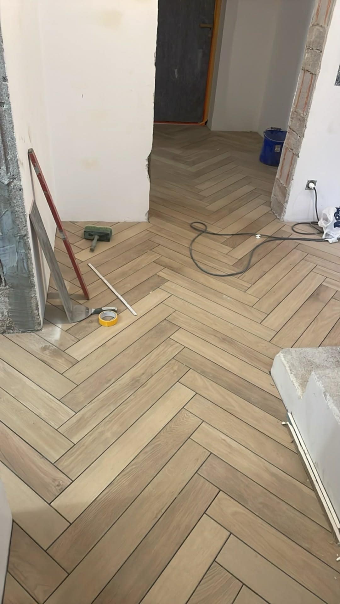 Ułożona podłoga z płytek w jodełkę, widoczne narzędzia glazurnicze: szpachelka, miarka, taśma. Jasne płytki imitujące drewno. Częściowo wykończone pomieszczenie.