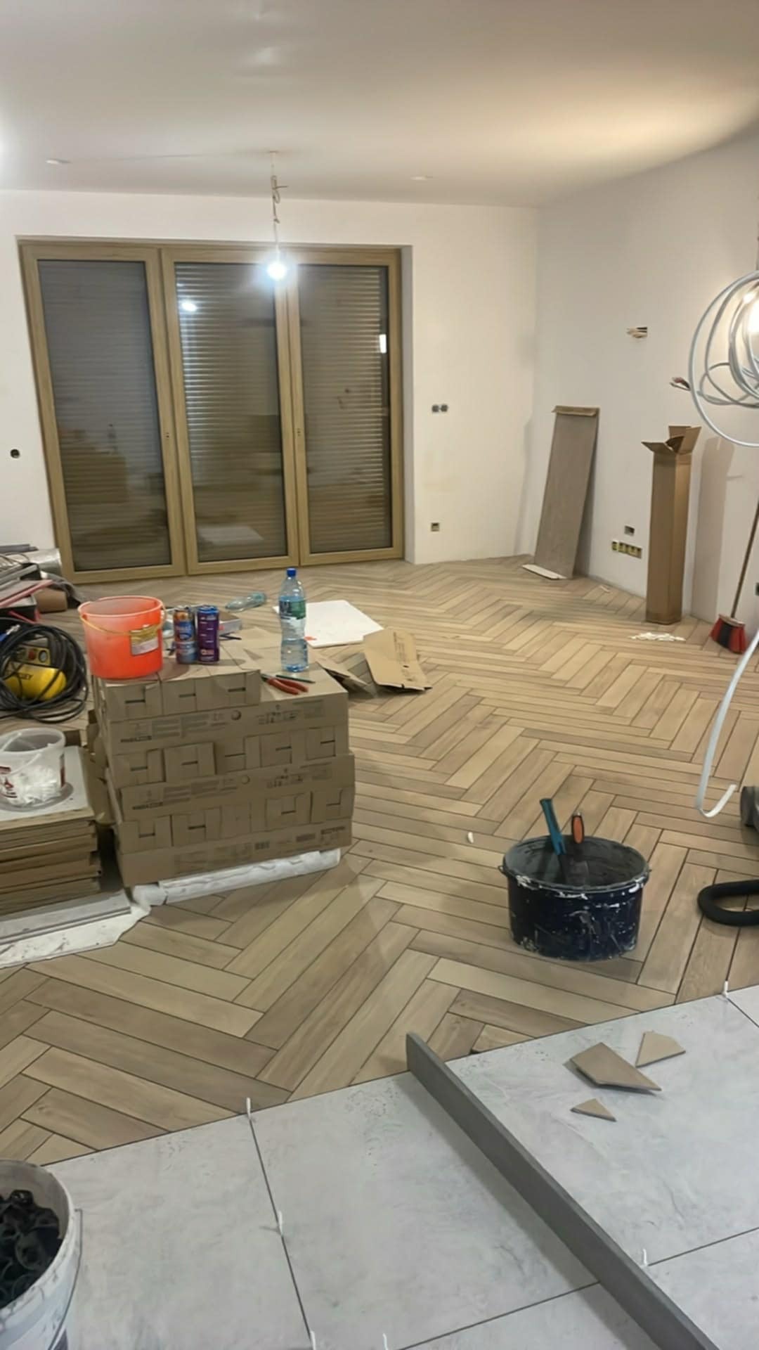 Jasne panele ułożone w jodełkę w przestronnym pokoju, częściowo zakryte kartonami i narzędziami. W tle duże okno balkonowe i białe ściany. Widoczny etap prac wykończeniowych.