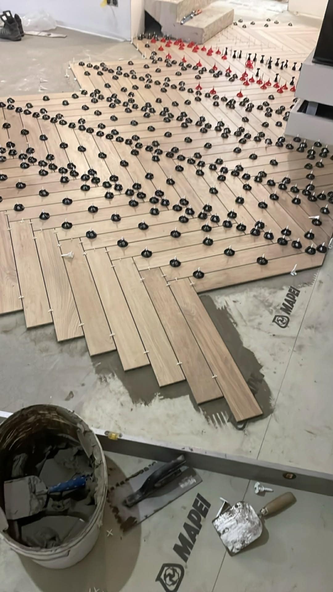 Układanie płytek imitujących drewno w jodełkę, z użyciem poziomujących klipsów i narzędzi glazurniczych. Widoczny kubeł z zaprawą i poziomica.