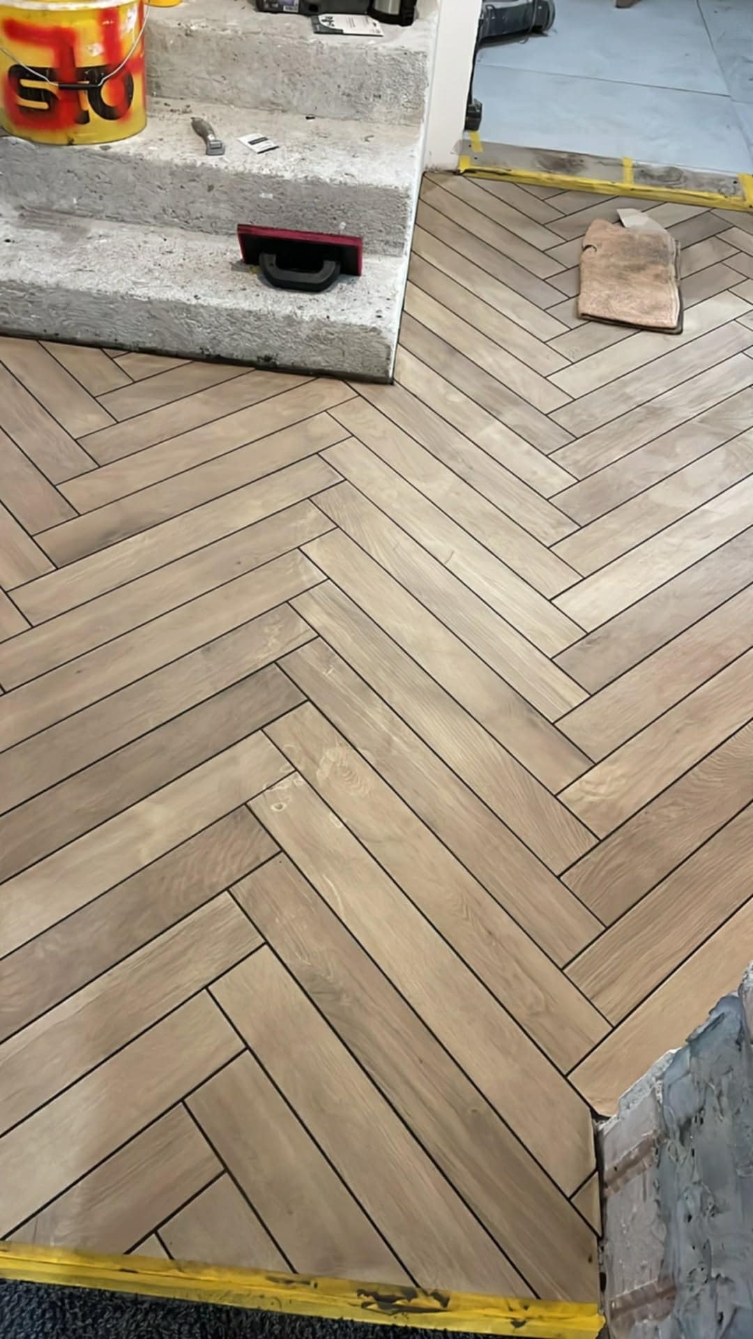 Ułożona w jodełkę podłoga z paneli imitujących drewno, widoczne narzędzia glazurnicze i fragment schodów. Jasne, naturalne światło podkreśla wzór.