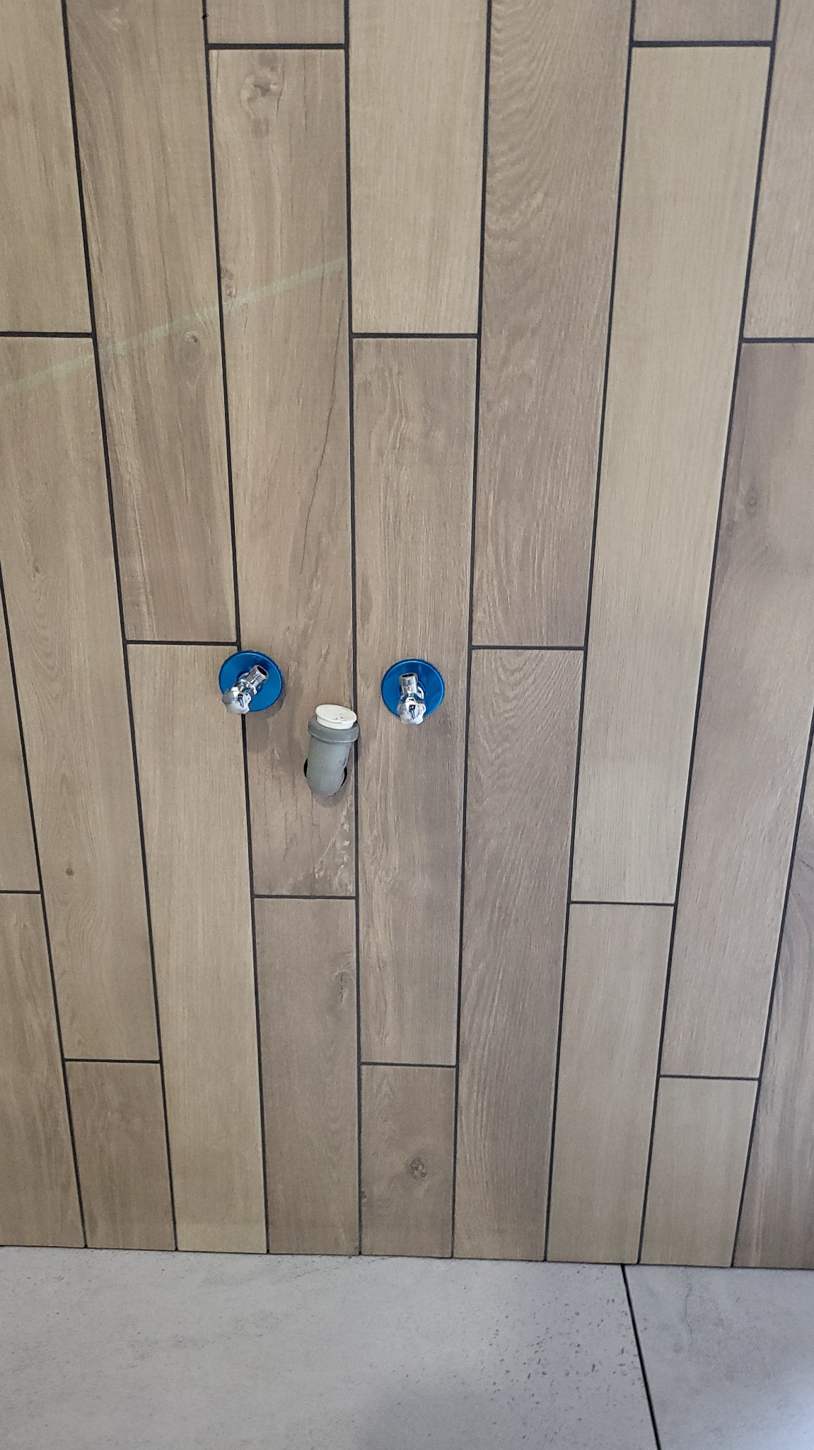 Ściana wyłożona podłużnymi, drewnopodobnymi płytkami z widocznymi przyłączami wodnymi i odpływem, ukazująca detale wykończenia w łazience.