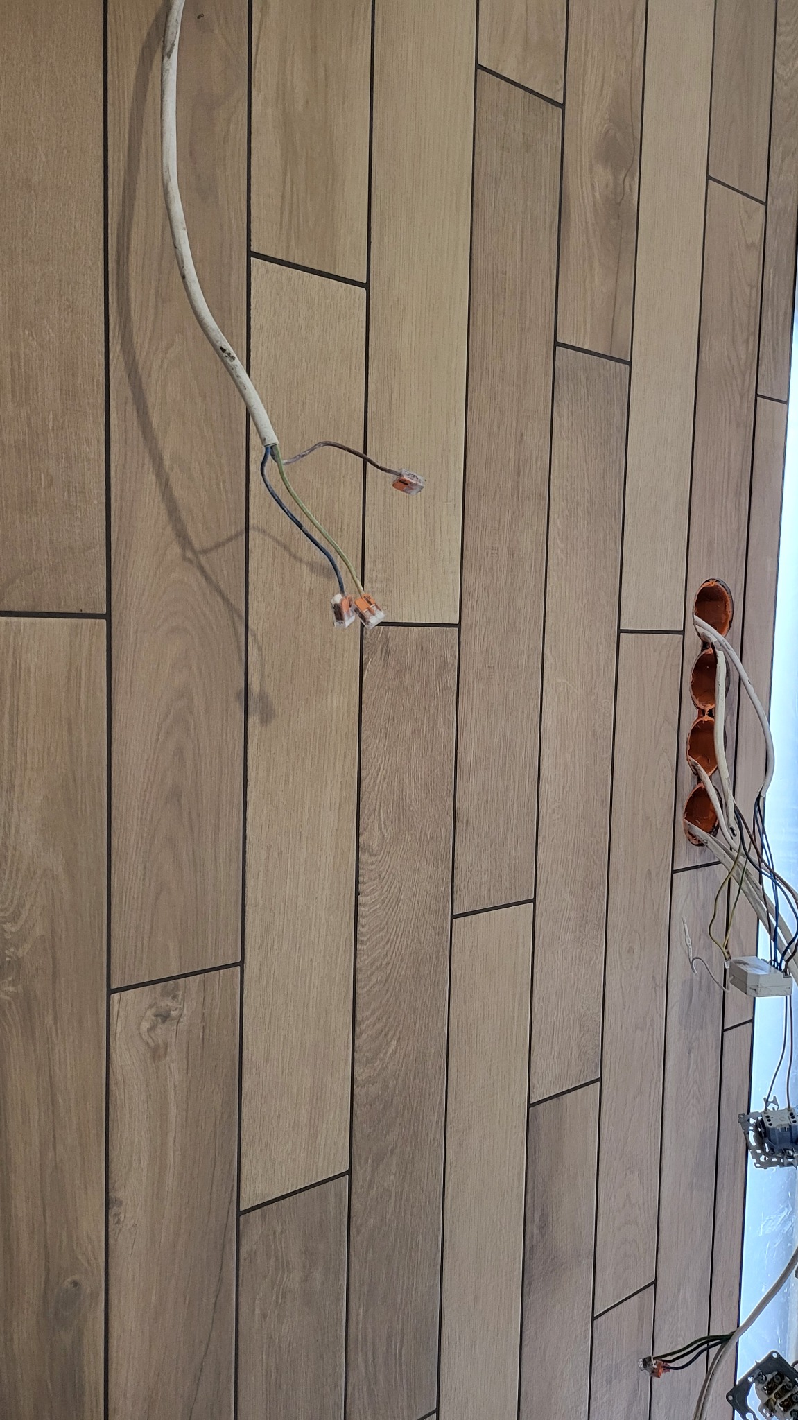 Ściana wyłożona podłużnymi płytkami imitującymi drewno, z widocznymi przewodami elektrycznymi i puszkami instalacyjnymi. Układ pionowy, wzór cegiełki.