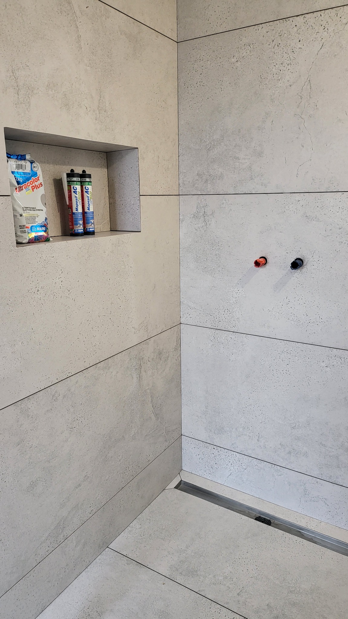 Wykończona ściana w łazience z szarych, matowych płytek imitujących kamień. Wnęka z chemią budowlaną i odpływ liniowy w podłodze. Widoczne przyłącza wodne.