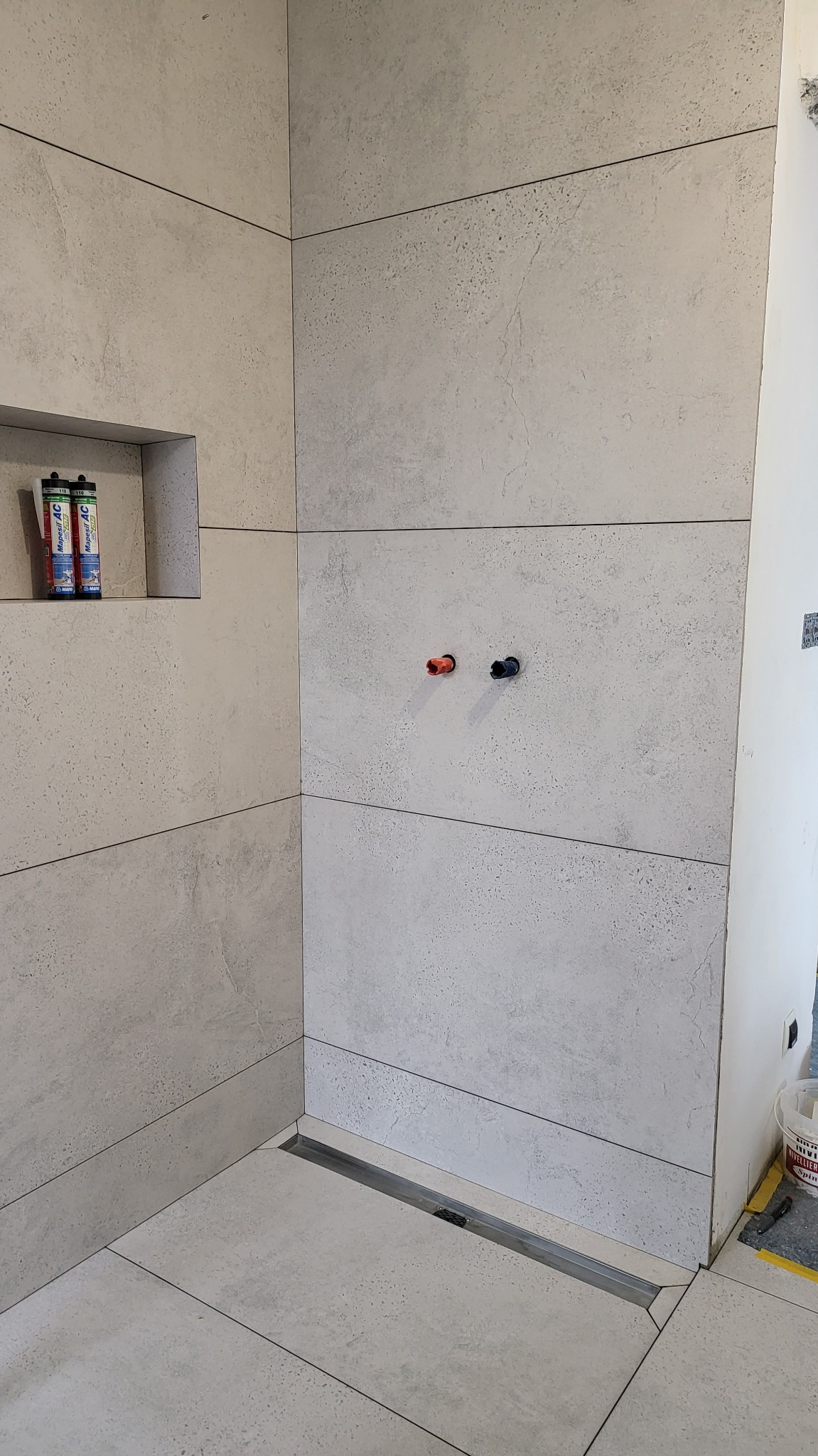 Wykończona kabina prysznicowa z szarymi, wielkoformatowymi płytkami, odpływem liniowym i wnęką z dwoma tubami uszczelniacza. Widoczne przyłącza wodne.