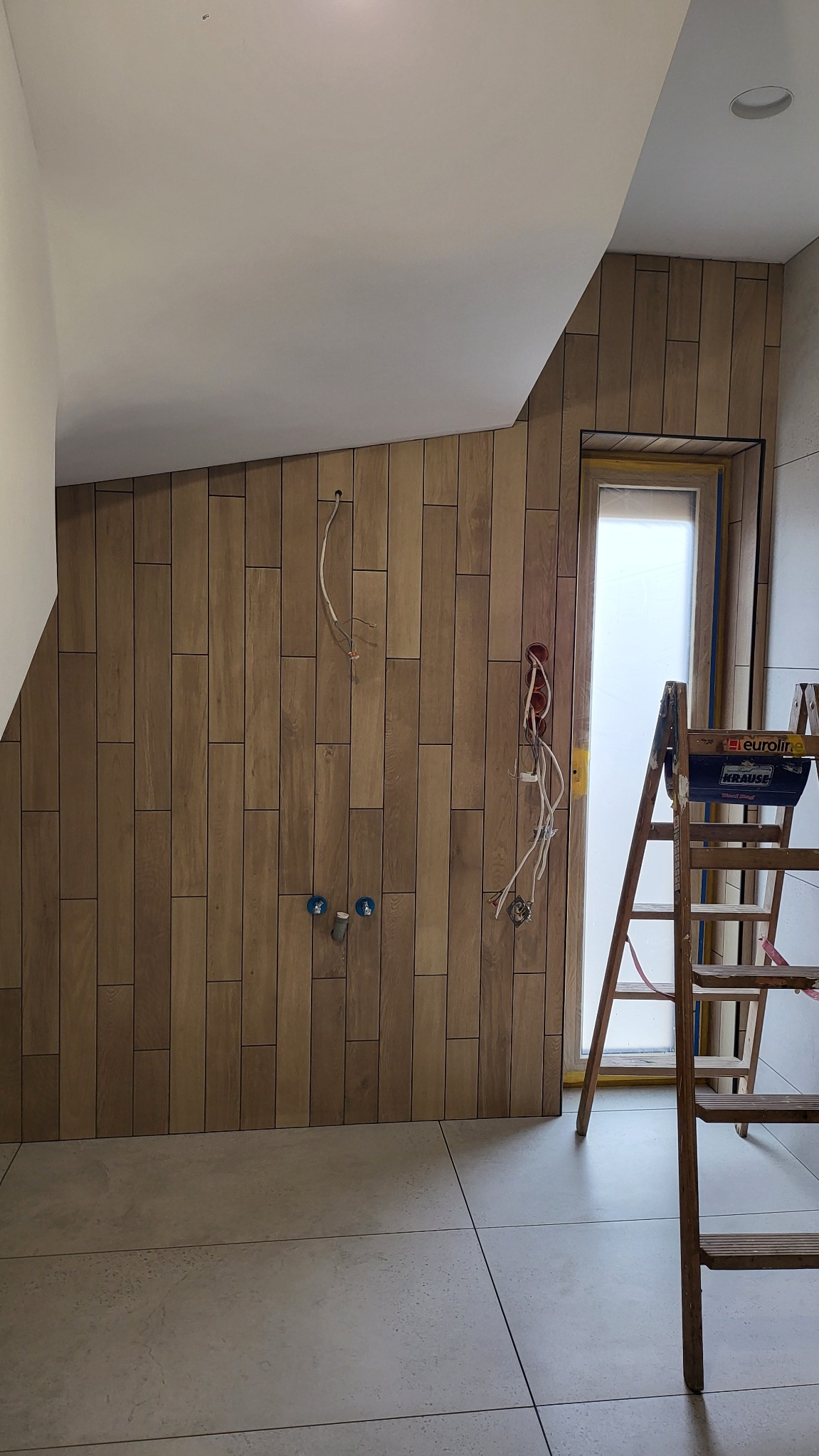 Ściana wyłożona drewnopodobnymi płytkami w łazience, widoczne wyprowadzenia hydrauliczne i elektryczne, drabina Krause. Ukończony etap prac.