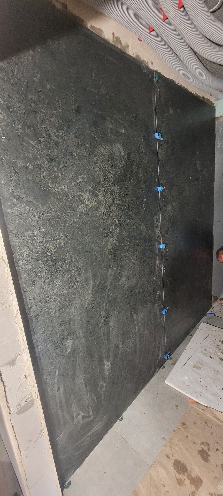 Układanie ciemnych, wielkoformatowych płytek na ścianie z użyciem poziomicy i krzyżyków dystansowych. Widoczne rury pod sufitem.