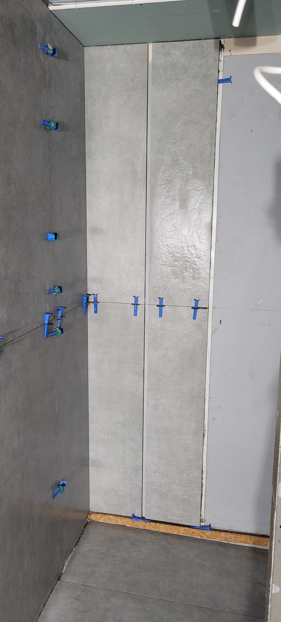 Układanie szarych płytek w Bydgoszczy z użyciem niebieskich krzyżyków dystansowych. Widok z góry na fragment ściany i podłogi w trakcie prac wykończeniowych.