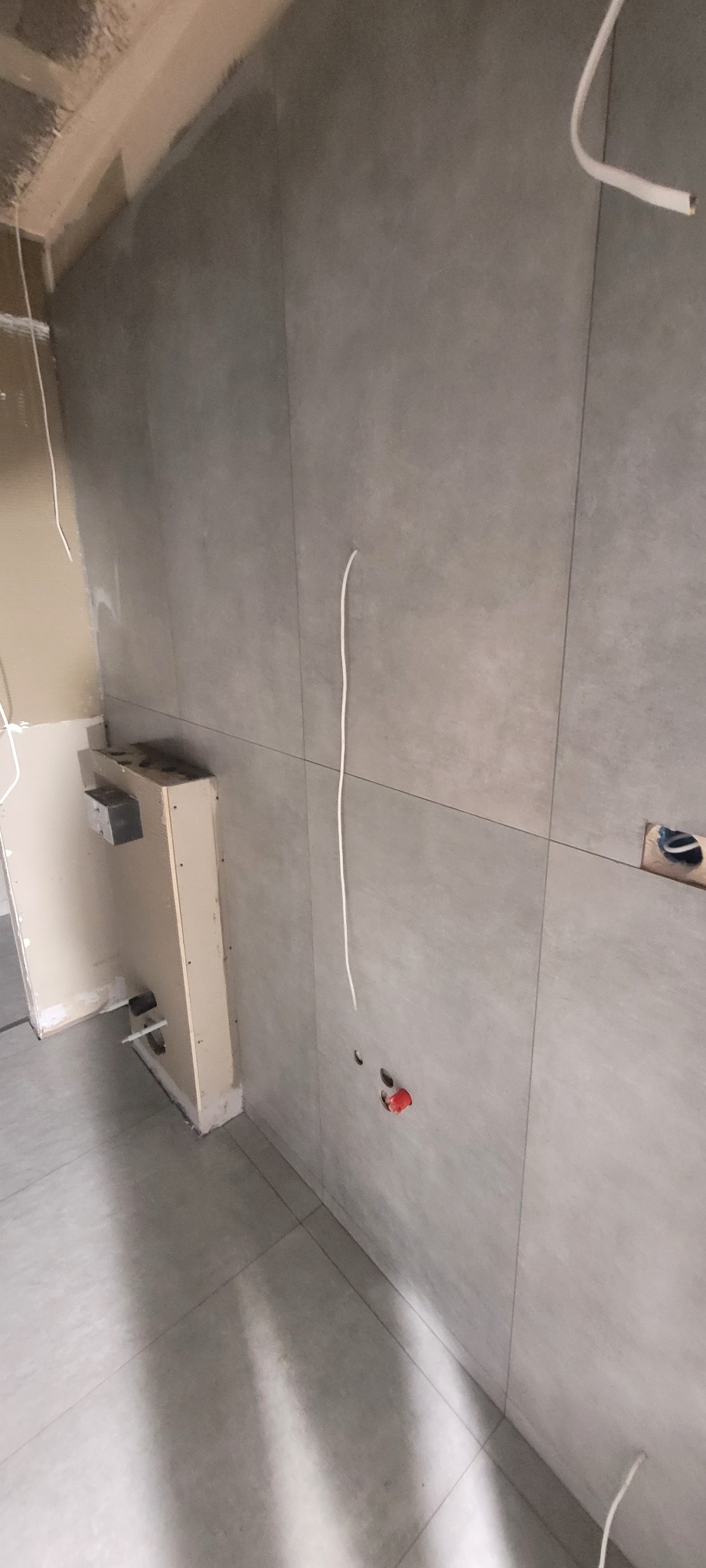 Ściana i podłoga wyłożone szarymi płytkami. Widoczne wystające kable i otwory w ścianie. Częściowo zabudowany element instalacji. Wnętrze w trakcie wykończenia.