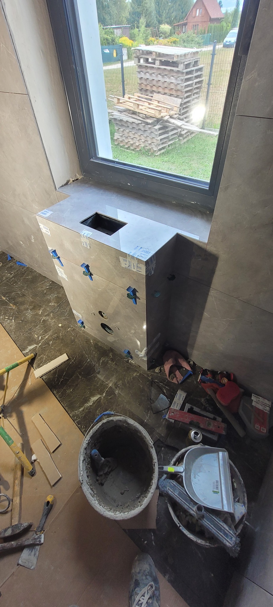 Wykończona obudowa umywalki z szarych płytek imitujących marmur, z niebieskimi krzyżykami dystansowymi, widok z góry na otwór na umywalkę, narzędzia glazurnicze w tle.