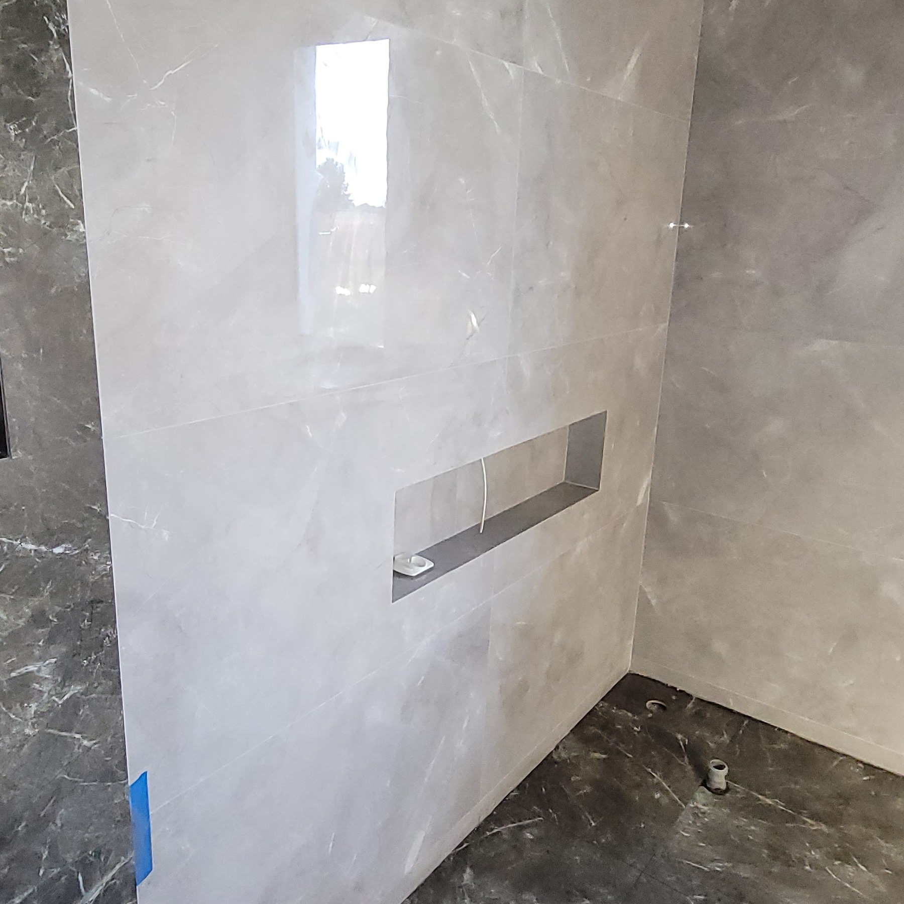 Wykończona łazienka z szarymi płytkami imitującymi marmur, wnęka w ścianie z półką. Widoczny fragment podłogi z ciemnymi płytkami. Minimalistyczny design.