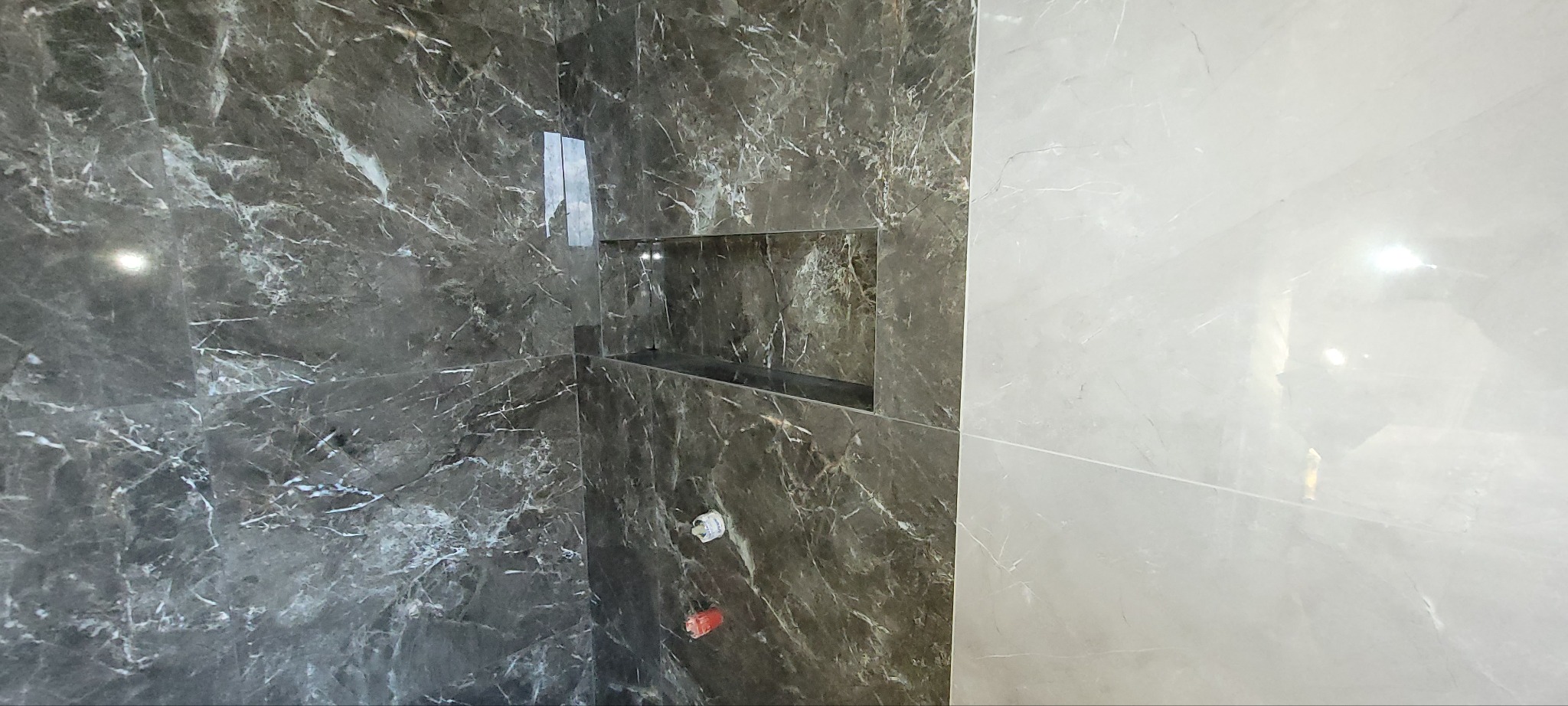 Wykończona wnęka w kabinie prysznicowej, obłożona płytkami imitującymi marmur w odcieniach szarości i bieli. Widoczne fugi i armatura.