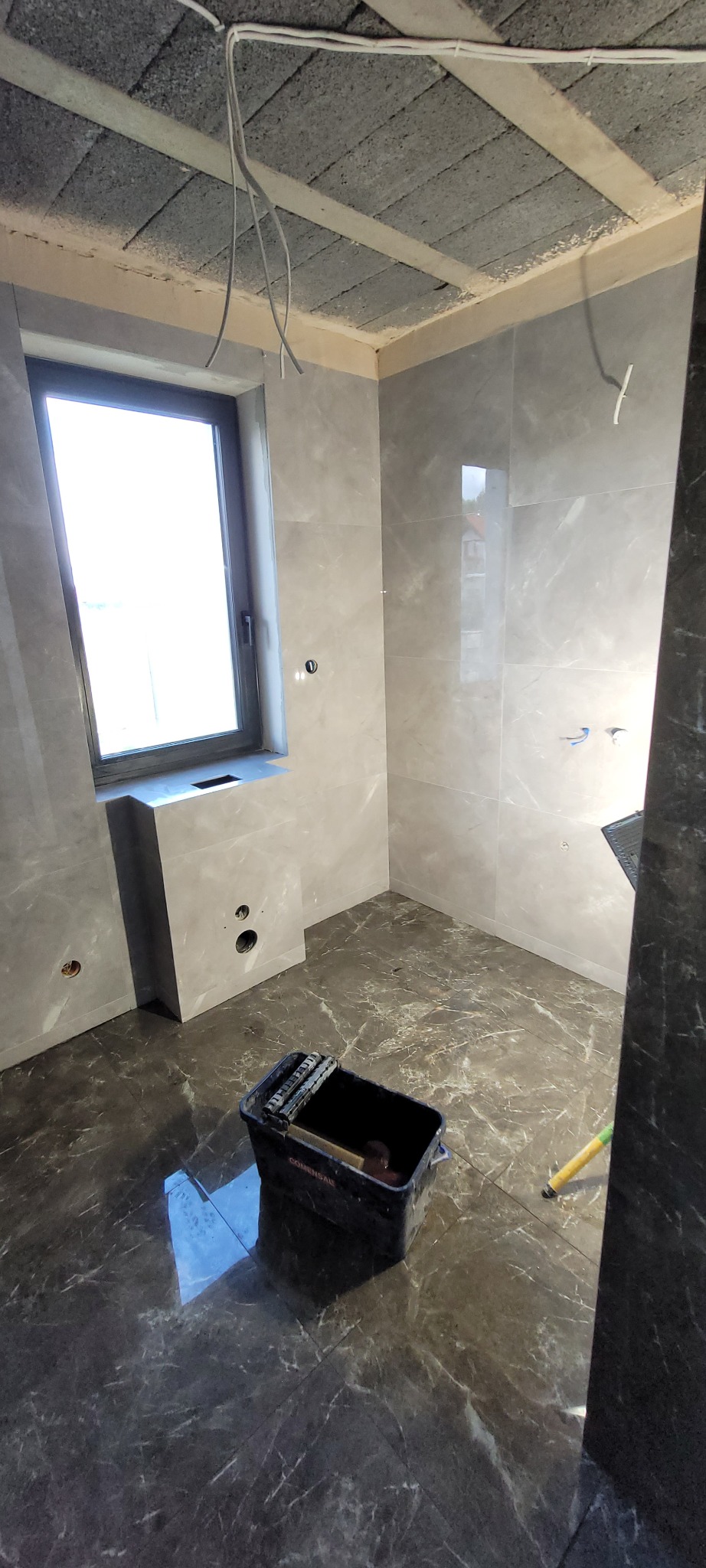 Wykończona łazienka z szarymi płytkami imitującymi marmur na ścianach i podłodze. Widoczny fragment okna i narzędzia glazurnicze.