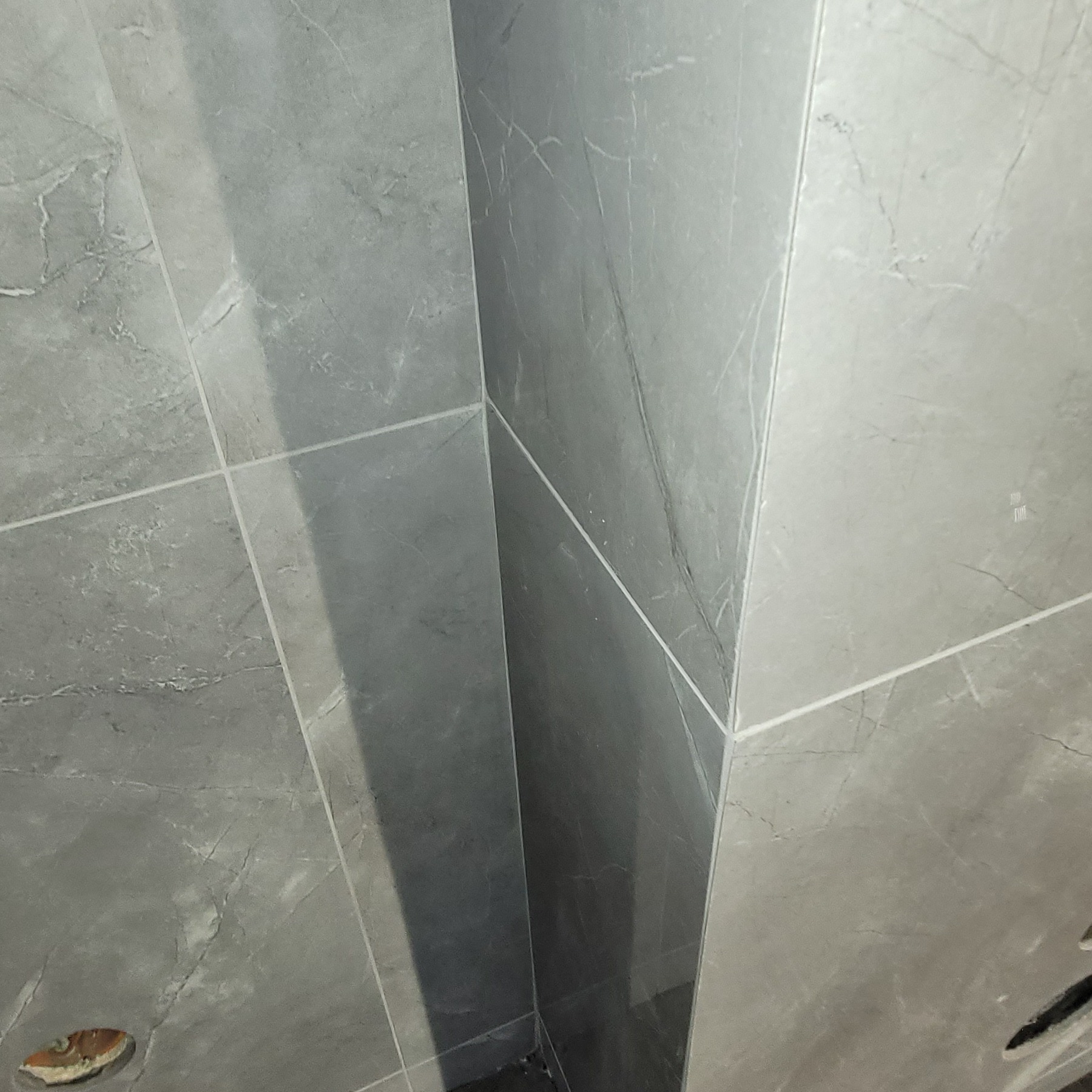 Wykończony narożnik łazienki z szarymi płytkami imitującymi marmur. Widoczne fugi i otwór na instalację. Minimalistyczny design.