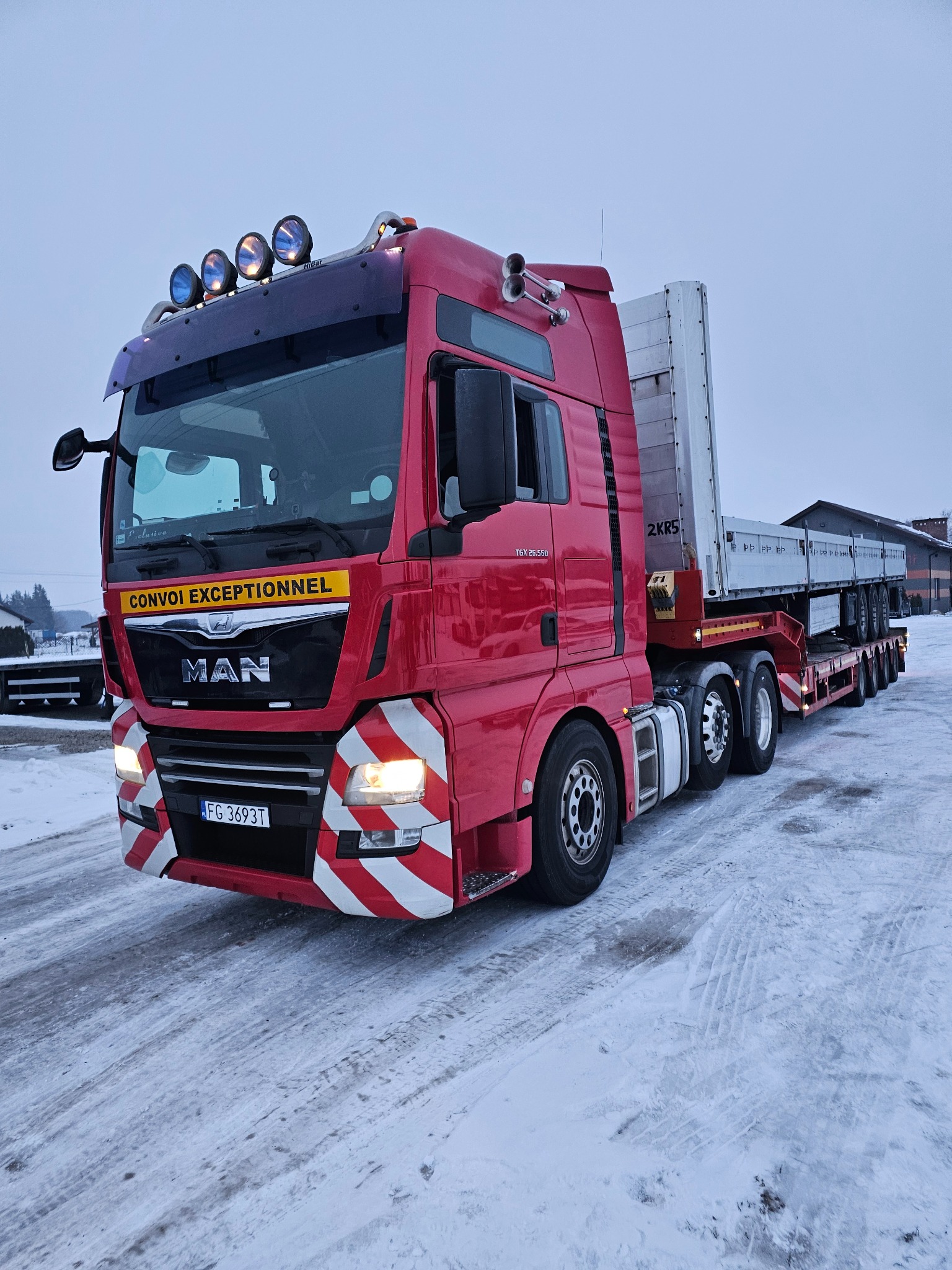 Czerwony ciągnik MAN z naczepą niskopodwoziową, oświetlenie robocze na dachu, tablica 'Convoi Exceptionnel', Stare Bielice. Transport ponadgabarytowy.