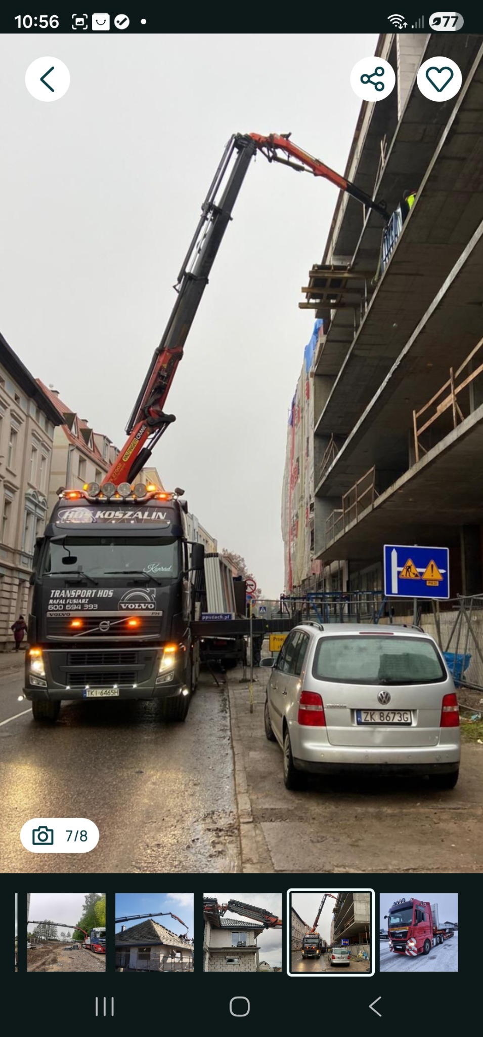 Ciężarówka HDS z wysięgnikiem podnosi materiały na budowę wielopiętrowego budynku w mieście. Na pierwszym planie zaparkowany srebrny samochód osobowy.