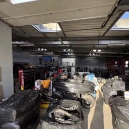 Mateusz Borkowski Trans Factory - Magazyn z zapakowanymi przedmiotami w czarną folię, przygotowanymi do transportu. Widoczne kartony, meble i inne przedmioty. Jasne światło wpada przez okna dachowe.