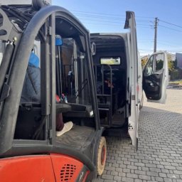 Mateusz Borkowski Trans Factory - Wózek widłowy Linde ładuje zabezpieczony ładunek do otwartego busa na brukowanym podjeździe, przygotowania do transportu.