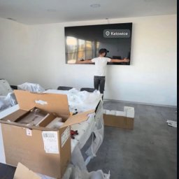 Mateusz Borkowski Trans Factory - Mężczyzna w czapce mierzy odległość na ścianie pod telewizorem w Katowicach. Kartony i folie ochronne sugerują przeprowadzkę lub montaż sprzętu.