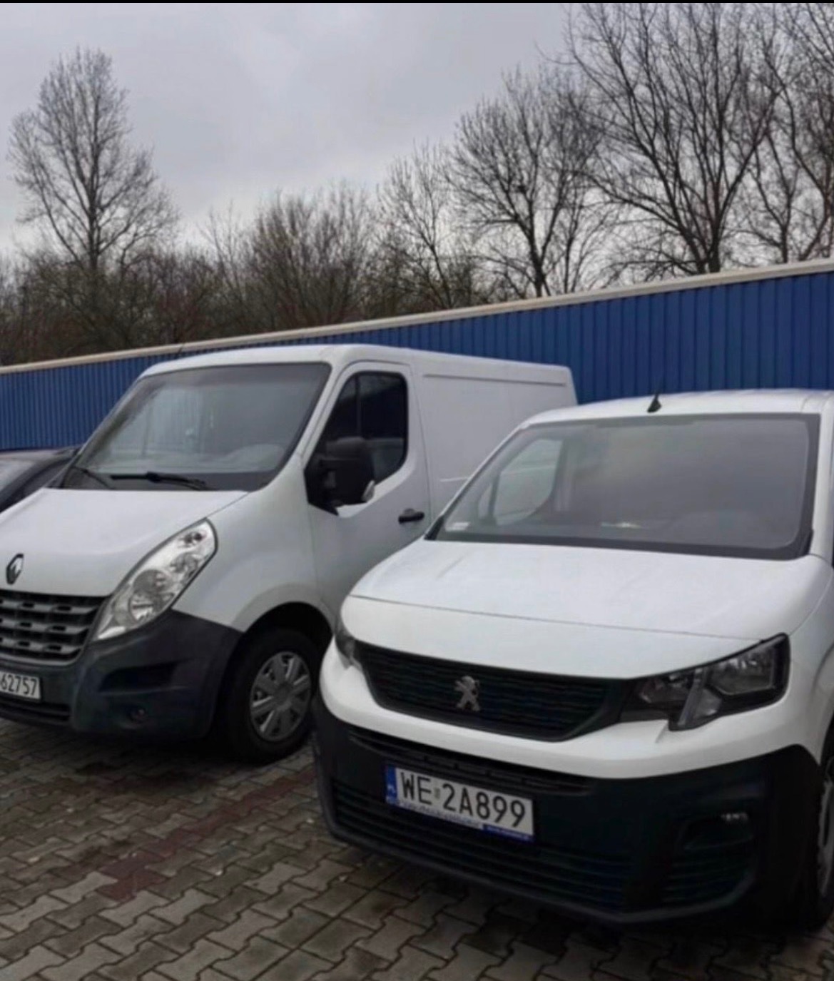 Dwa białe busy: Renault Master z widoczną tablicą rejestracyjną i Peugeot Partner, zaparkowane przed niebieskim magazynem. Pochmurne niebo w tle.