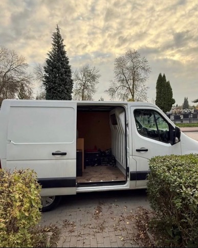 Otwarty biały furgon na tle drzew i pochmurnego nieba, częściowo załadowany meblami, sugerujący gotowość do transportu. Widok z boku, krzewy w pierwszym planie.