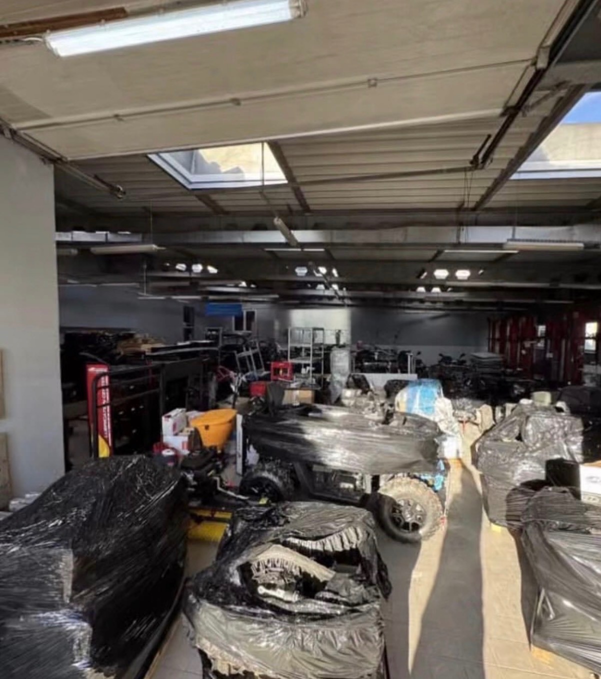 Magazyn z zapakowanymi przedmiotami w czarną folię, przygotowanymi do transportu. Widoczne kartony, meble i inne przedmioty. Jasne światło wpada przez okna dachowe.