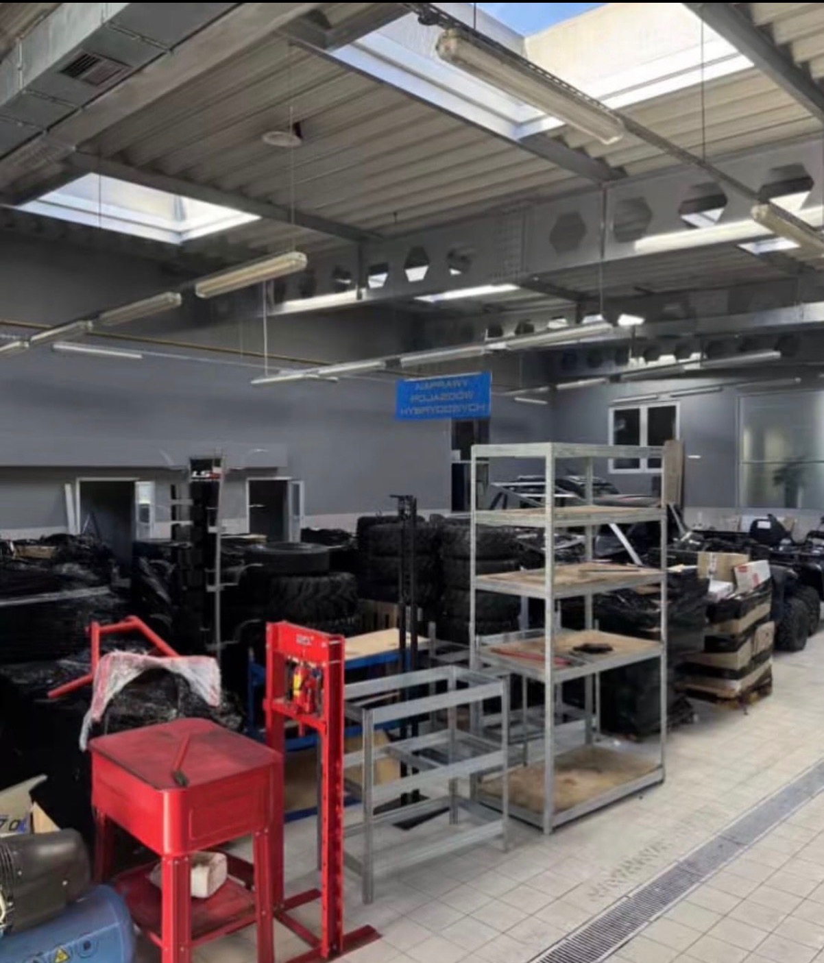 Magazyn z oponami, regałami i narzędziami w hali. Widoczne opony, metalowe regały, czerwony stół warsztatowy i niebieski kompresor. Hala z oknami w suficie.