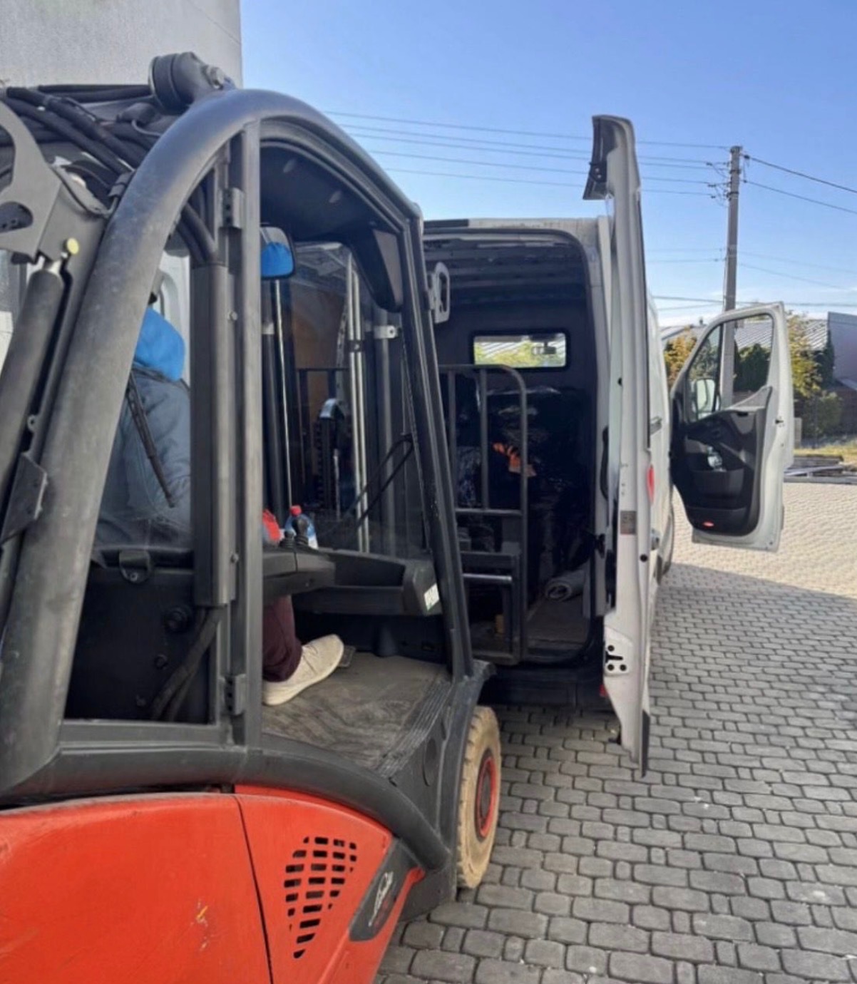 Wózek widłowy Linde ładuje zabezpieczony ładunek do otwartego busa na brukowanym podjeździe, przygotowania do transportu.