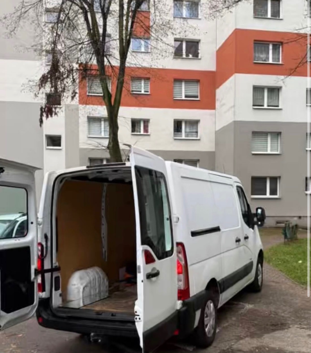 Otwarty, pusty furgon dostawczy na tle bloku mieszkalnego, gotowy do załadunku. Widoczne wnętrze paki i fragment chodnika.