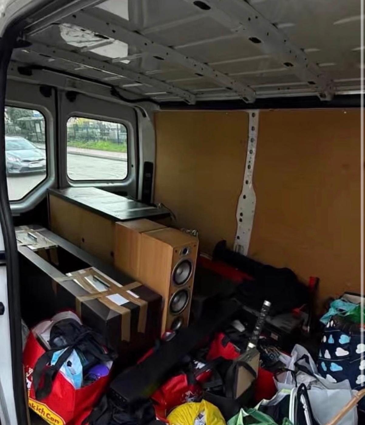 Wnętrze furgonetki załadowane sprzętem audio, torbami i kartonami, przygotowane do transportu. Widoczne głośniki i inne przedmioty zabezpieczone na czas przewozu.