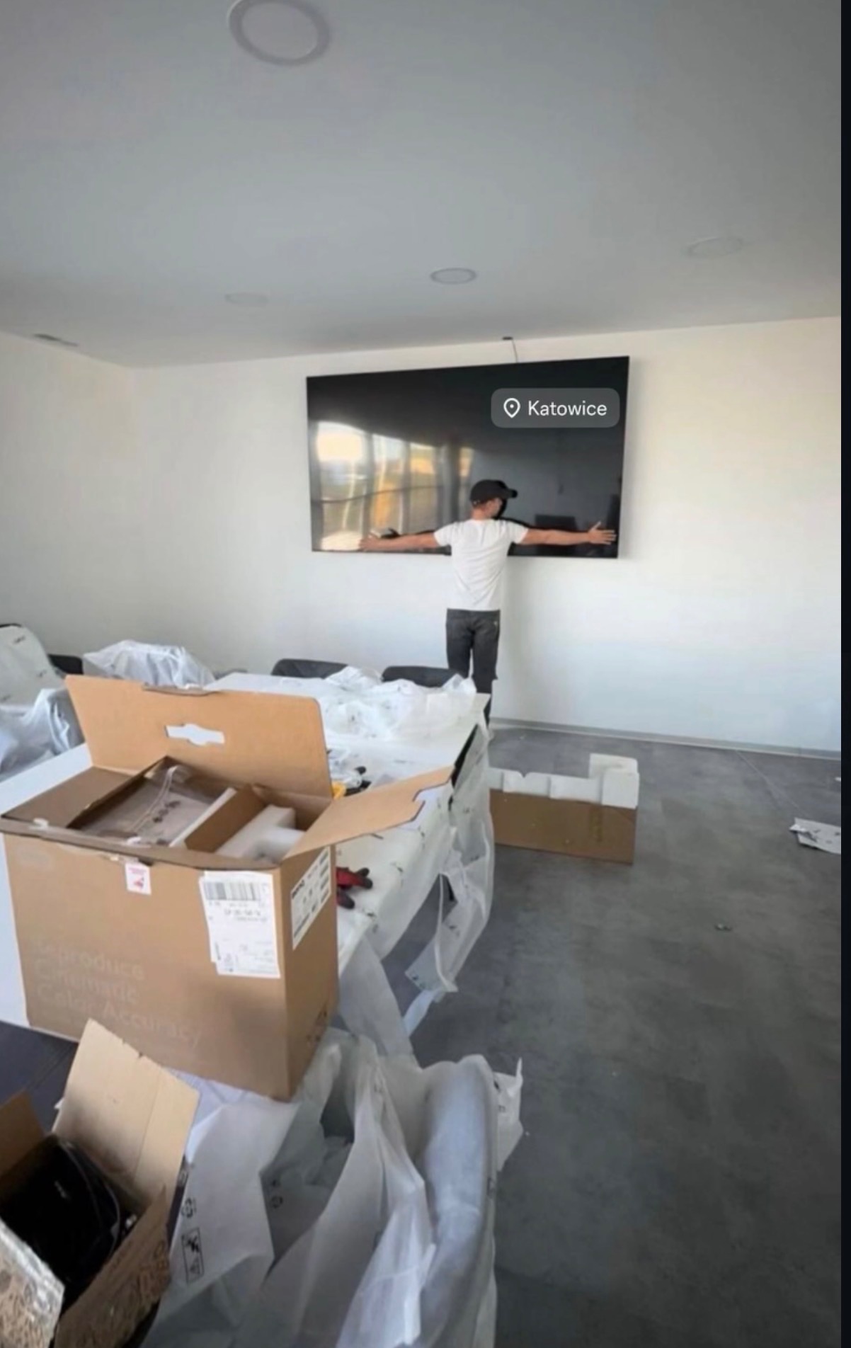 Mężczyzna w czapce mierzy odległość na ścianie pod telewizorem w Katowicach. Kartony i folie ochronne sugerują przeprowadzkę lub montaż sprzętu.