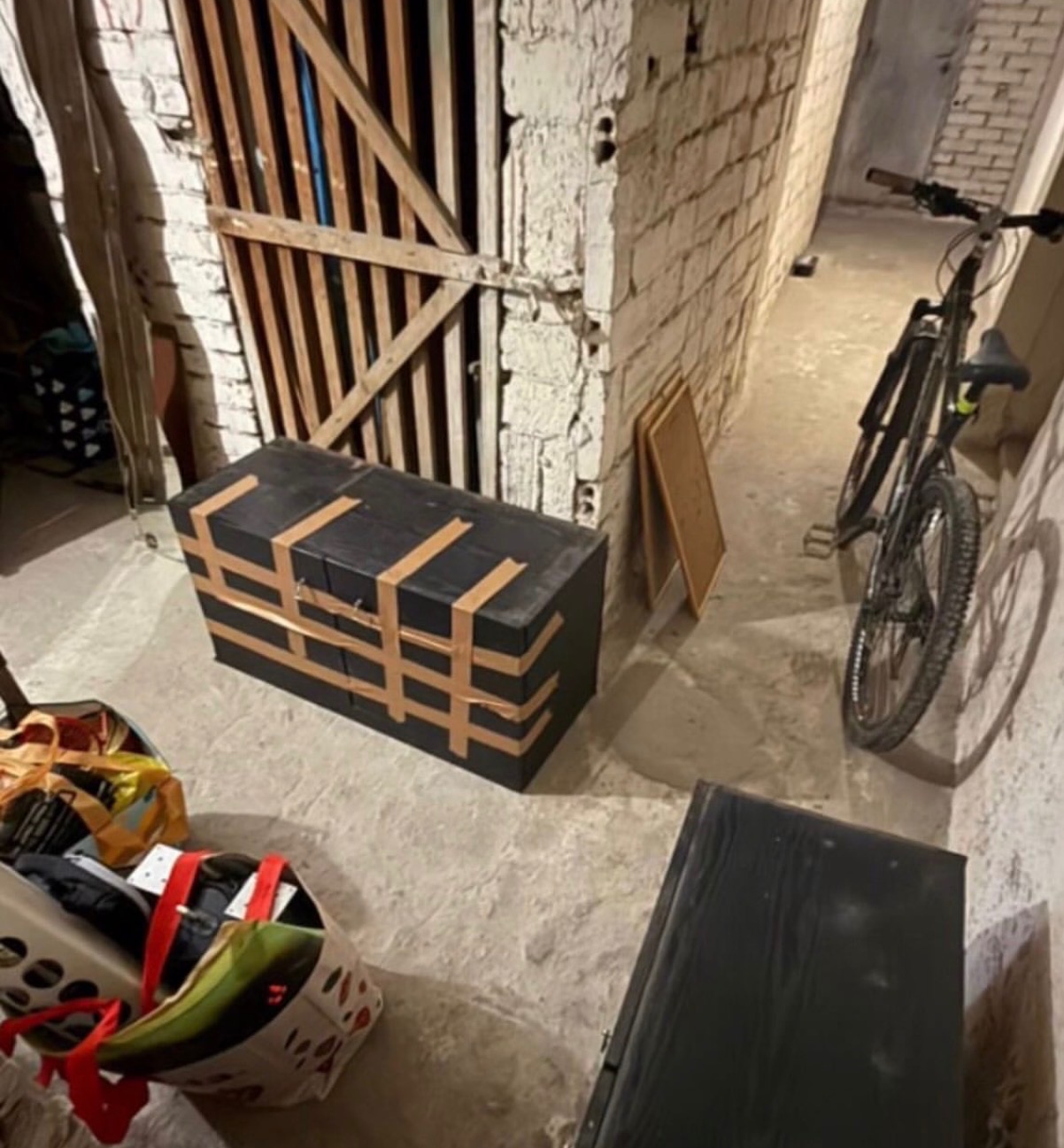 Spakowany karton oklejony taśmą, gotowy do transportu w piwnicy. Obok rower i torby z rzeczami, ściana z cegły i drewniane drzwi.