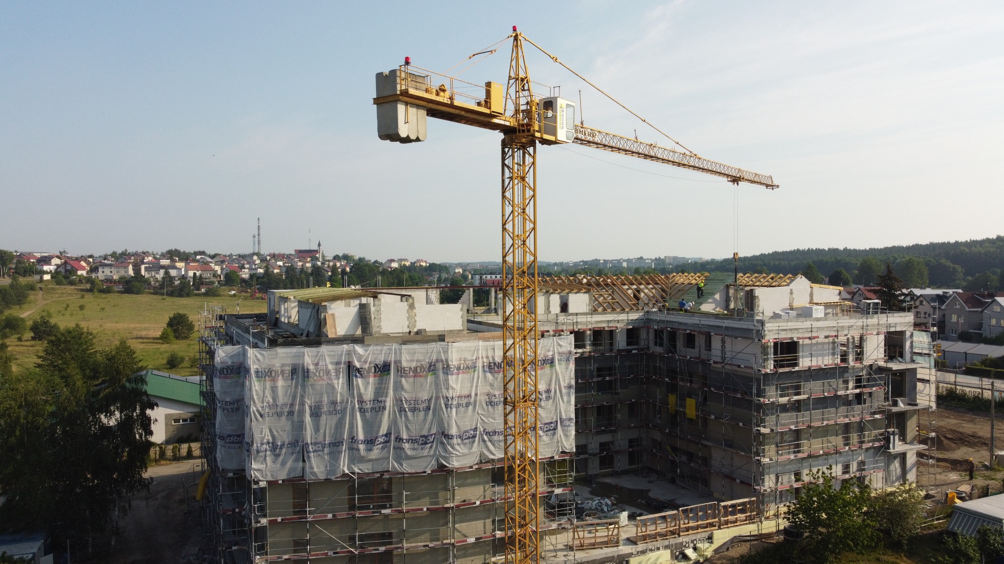 Żuraw budowlany na tle wznoszonego budynku wielorodzinnego z betonu komórkowego, osłoniętego siatką ochronną z reklamami. W tle zabudowa miejska i zieleń.