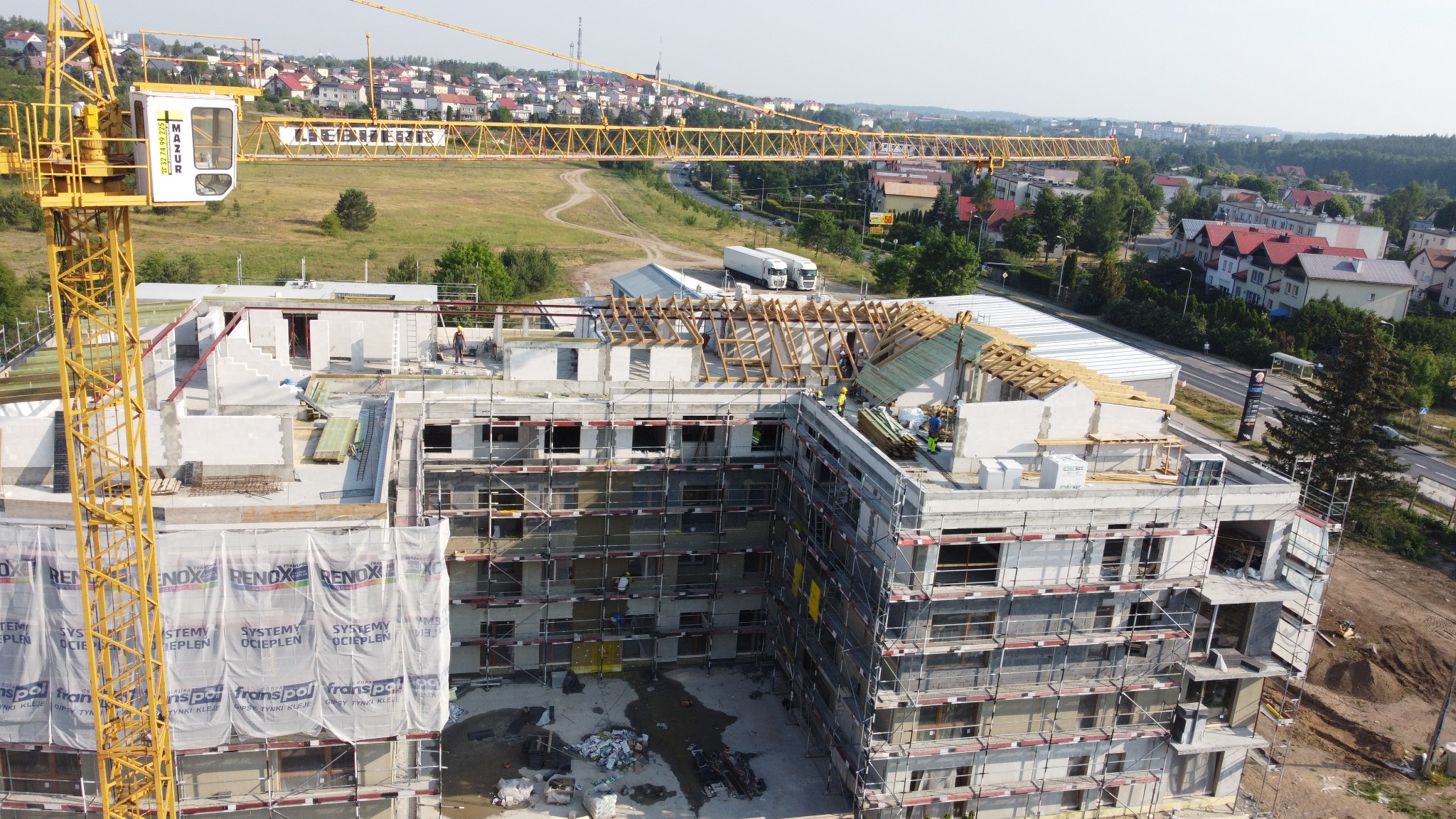 Budowa budynku żelbetowego w toku, widoczne rusztowania, dźwig Liebherr, częściowo zadaszona konstrukcja, materiały budowlane i pracownicy w kaskach. Ujęcie z góry.