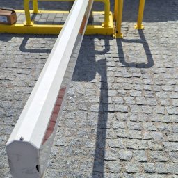 IT 4.0 SPÓŁKA Z OGRANICZONĄ ODPOWIEDZIALNOŚCIĄ - Szlaban automatyczny z żółtymi barierkami ochronnymi na kostce brukowej, częściowo zasłonięty przez zaparkowany samochód. Jasne, naturalne oświetlenie.