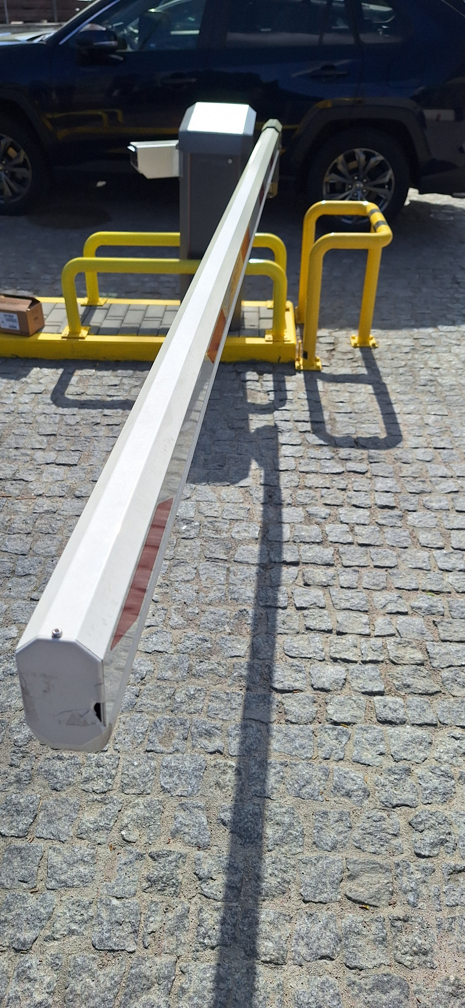 Szlaban automatyczny z żółtymi barierkami ochronnymi na kostce brukowej, częściowo zasłonięty przez zaparkowany samochód. Jasne, naturalne oświetlenie.