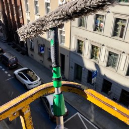 ALLCLEAN Usługi Sprzątające JOACHIM SCHÄFER - Szczotka do mycia okien na wysięgniku, widok z góry na ulicę miejską z budynkami w tle. Żółta barierka w kadrze. Słoneczny dzień.