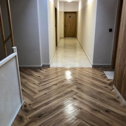 ALLCLEAN Usługi Sprzątające JOACHIM SCHÄFER - Wnętrze korytarza po sprzątaniu: parkiet ułożony w jodełkę, jasne ściany z szarym pasem u góry, czyste płytki w dalszej części korytarza, widoczne drzwi.