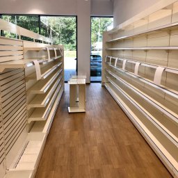 ALLCLEAN Usługi Sprzątające JOACHIM SCHÄFER - Pusta przestrzeń handlowa z drewnianą podłogą i białymi regałami, widok na zewnątrz przez okno. Przygotowanie do otwarcia sklepu lub po sprzątaniu.