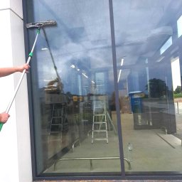 ALLCLEAN Usługi Sprzątające JOACHIM SCHÄFER - Mycie dużej szyby okiennej z użyciem mopa na teleskopowym kiju. Widoczna drabina i otoczenie budynku. Szyba częściowo umyta, widać różnicę w czystości.