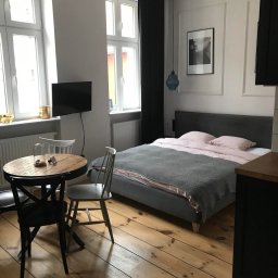 ALLCLEAN Usługi Sprzątające JOACHIM SCHÄFER - Jasne wnętrze sypialni z drewnianą podłogą, okrągłym stolikiem i łóżkiem z szarą narzutą. Minimalistyczny wystrój z czarnymi akcentami i naturalnym światłem.