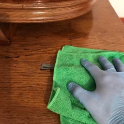 ALLCLEAN Usługi Sprzątające JOACHIM SCHÄFER - Ręka w rękawicy lateksowej czyści drewniany blat zieloną ściereczką z mikrofibry marki Karcher. Widoczny fragment blatu stołu z ciemnego drewna.