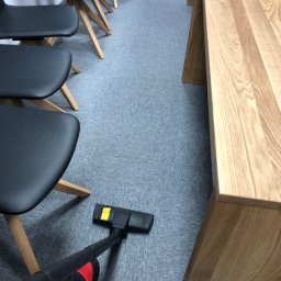 ALLCLEAN Usługi Sprzątające JOACHIM SCHÄFER - Szary dywan w biurze odkurzany odkurzaczem z czarną rurą i żółtymi akcentami, obok drewniany stół i czarne krzesła na drewnianych nogach.