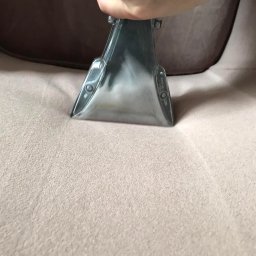 ALLCLEAN Usługi Sprzątające JOACHIM SCHÄFER - Dysza ekstraktora w trakcie prania jasnej, tekstylnej tapicerki kanapy. Widoczna ręka operatora i fragment oparcia. Delikatne oświetlenie.