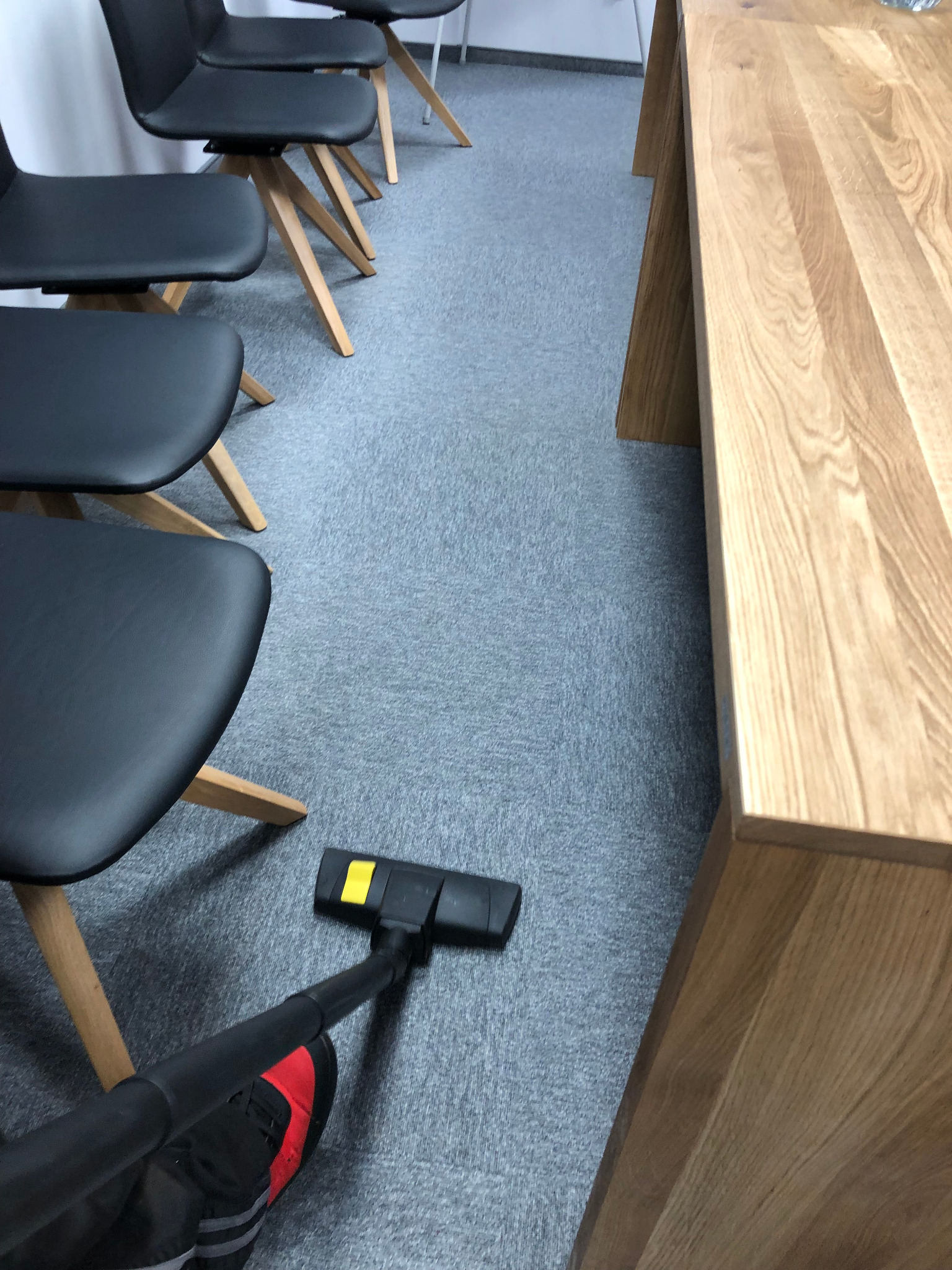 Szary dywan w biurze odkurzany odkurzaczem z czarną rurą i żółtymi akcentami, obok drewniany stół i czarne krzesła na drewnianych nogach.