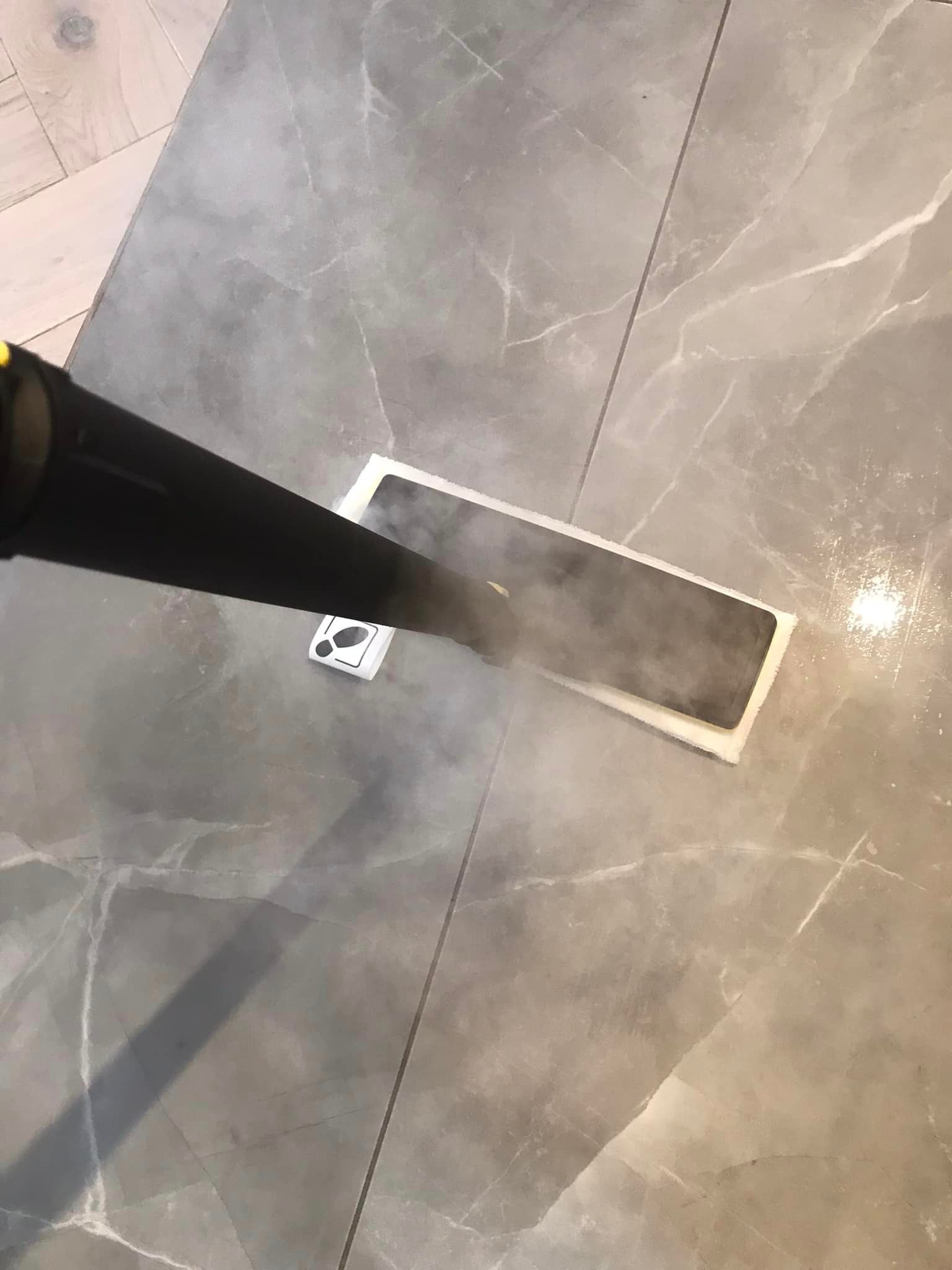 Czyszczenie parowe szarych płytek podłogowych z wzorem marmuru za pomocą czarnego mopa parowego. Widoczna para i naklejka z symbolem buta.