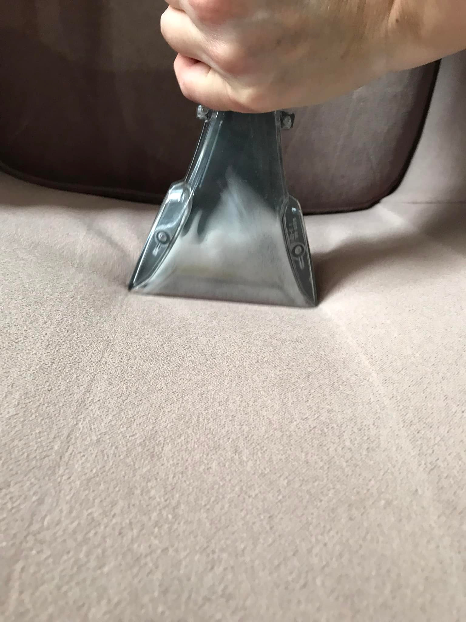 Dysza ekstraktora w trakcie prania jasnej, tekstylnej tapicerki kanapy. Widoczna ręka operatora i fragment oparcia. Delikatne oświetlenie.